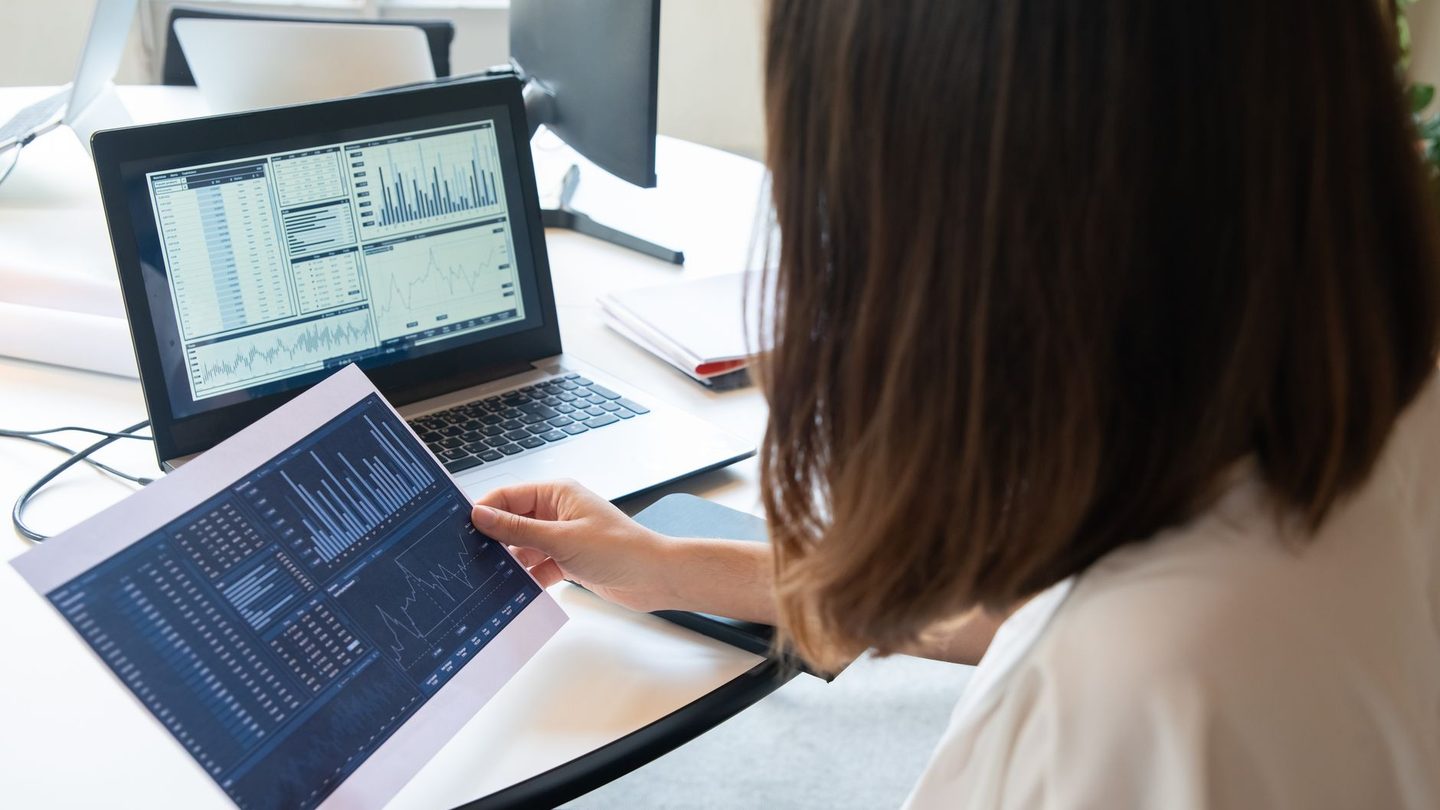 An analyst at a desk holding a printed page of tables beside a laptop showing more tables